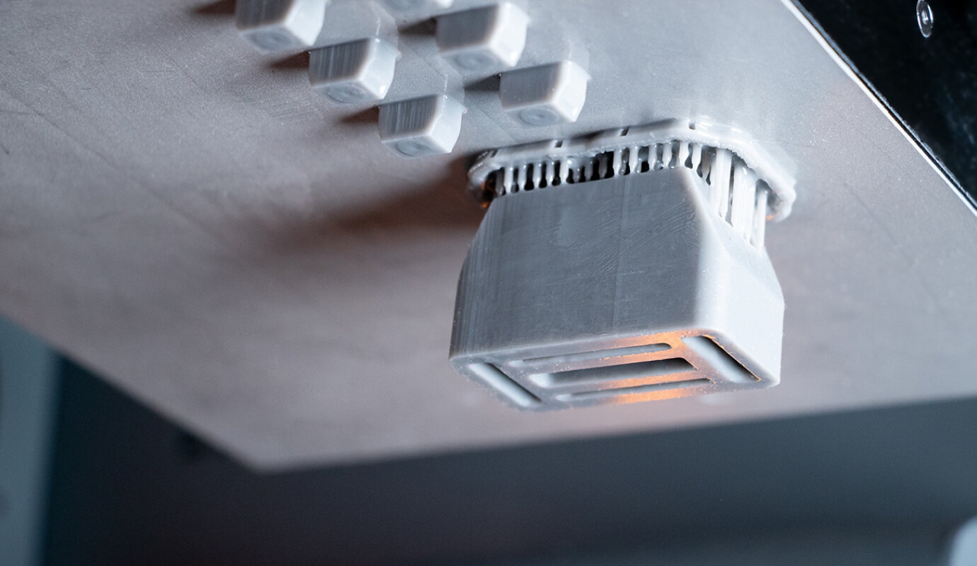 Nahaufnahme eines 3D-Druckprozesses im FDM-Verfahren, bei dem ein graues Kunststoffbauteil kopfüber mit sichtbaren Stützstrukturen an der Druckplatte haftet. Die feinen Drucklinien und schichtweise Aufbauweise des Objekts sind gut erkennbar.