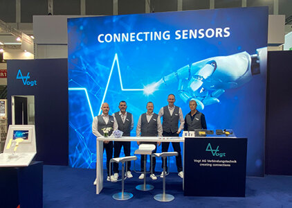 Messestand der Vogt AG Verbindungstechnik mit fünf Mitarbeitenden in einheitlicher Kleidung vor einer großflächigen, blau beleuchteten Wand mit dem Slogan „Connecting Sensors“ und einer Grafik eines Roboterarms. Im Vordergrund befinden sich ein Stehtisch mit Hockern sowie ein Empfangstresen mit Firmenlogo.