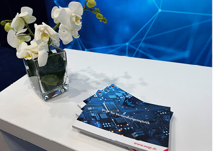 Nahaufnahme eines weißen Messetresens mit einem quadratischen Glasgefäß mit weißen Orchideen und mehreren Broschüren der Vogt AG Verbindungstechnik, die auf dem Tisch ausgelegt sind. Im Hintergrund ist eine blaue Wand mit abstraktem, technologischem Linienmuster sichtbar.