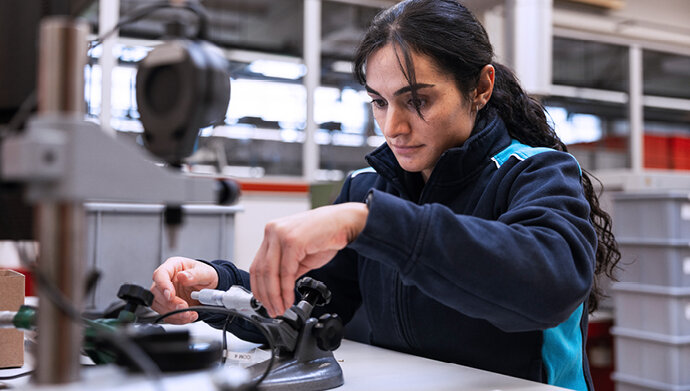 Eine Mitarbeiterin führt in einer Fertigungshalle eine manuelle Qualitätsprüfung durch, indem sie ein Bauteil in einer Messvorrichtung kontrolliert. Ihre konzentrierte Haltung unterstreicht die Präzision des Prozesses.