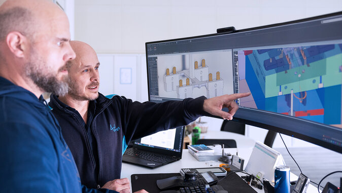 Zwei Ingenieure betrachten ein CAD-Modell auf einem großen, gebogenen Monitor, während einer auf eine komplexe 3D-Bauteildarstellung zeigt. Die Darstellung zeigt verschiedene Ansichten mit Einsätzen und Aussparungen, vermutlich im Rahmen einer Design-for-Manufacturing-Prüfung.