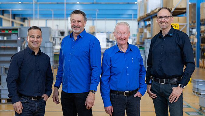 Vier Männer, Mitglieder der Geschäftsleitung, stehen lächelnd in einer hell beleuchteten Industrie- oder Produktionshalle. Sie tragen alle dunkle Jeans und blaue Hemden, was ihnen ein einheitliches und professionelles Auftreten verleiht.
