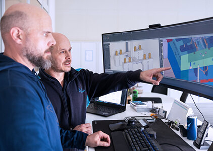 Zwei Männer stehen an einem Arbeitsplatz und betrachten einen großen, gebogenen Monitor, auf dem 3D-CAD-Modelle angezeigt werden; einer von ihnen zeigt auf den Bildschirm und erklärt offenbar Details. Die Szene vermittelt eine technische Zusammenarbeit im Bereich Konstruktion oder Produktentwicklung mit digitaler Modellierungssoftware.