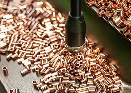 Zahlreiche kleine Kupferhülsen liegen auf einem Förderband oder einer Metalloberfläche, während ein automatisierter Greifer oder Sensor über ihnen positioniert ist. Die Szene stammt aus einem Glühofen-Prozess zur Wärmebehandlung der Hülsen.