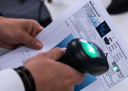 Eine Person scannt mit einem Handscanner einen Barcode auf einem Prüfprotokoll der Firma Vogt. Das Dokument enthält technische Informationen und eine Abbildung, was auf einen Qualitätssicherungsprozess in der Produktion hinweist.