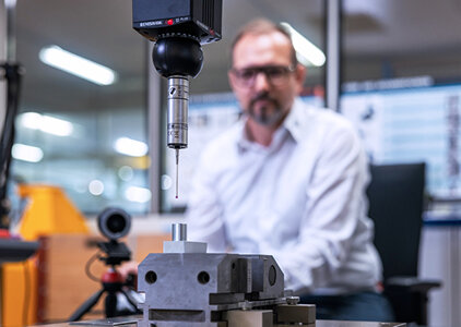 Im Vordergrund misst ein 3D-Taster eines Koordinatenmessgeräts präzise ein Metallbauteil, das in einer Spannvorrichtung fixiert ist. Im Hintergrund beobachtet ein Mann in Hemd und Brille konzentriert den Messvorgang – typisch für die Qualitätssicherung in der Fertigung.