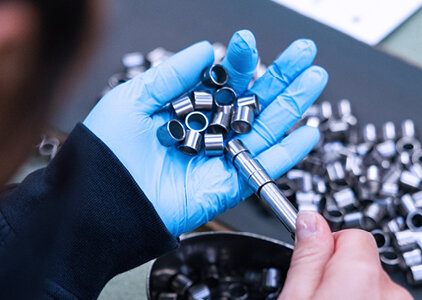 Nahaufnahme einer Hand mit blauem Handschuh, die mehrere kleine Metallhülsen hält, während mit einem Messgerät die Maße kontrolliert werden. Die Szene zeigt eine präzise Qualitätskontrolle in der industriellen Fertigung.