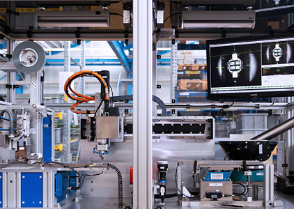 Blick auf eine vollautomatisierte Prüf- und Montagezelle mit Roboterarmen, Fördertechnik und einem Monitor, der das Prüfergebnis eines Bauteils zeigt. Die Anlage befindet sich in einer modernen Produktionshalle und dient der präzisen Qualitätskontrolle.