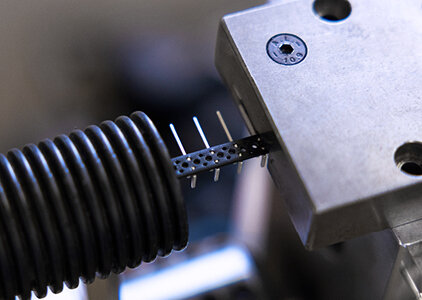 Makroaufnahme eines Steckergehäuses mit mehreren herausragenden Metallkontakten, das in eine Spannvorrichtung eingespannt ist. Die Szene zeigt einen präzisen Montageschritt oder eine Kontaktierungsprüfung in der Kabelkonfektionierung.