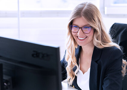 Eine lächelnde Mitarbeiterin mit Brille sitzt vor einem Computerbildschirm und wirkt engagiert im Kundengespräch oder bei der Büroarbeit. Die helle Büroumgebung unterstreicht die professionelle, aber freundliche Atmosphäre.