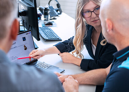 Eine Beraterin erklärt zwei Kunden Details auf einem Laptop-Bildschirm und zeigt mit einem Stift auf eine technische Zeichnung. Die Szene findet in einem modernen Büro statt und vermittelt professionelle Kundenberatung.