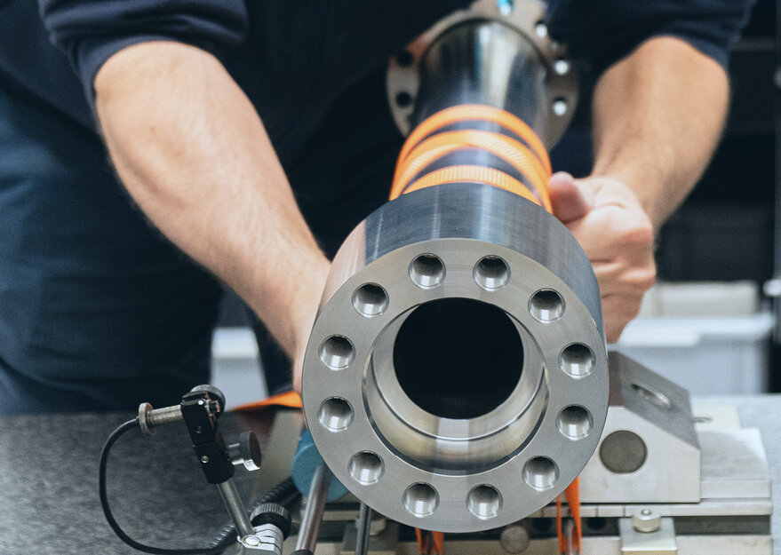 Nahaufnahme einer Prüfsituation im Messlabor, bei der eine Person ein zylindrisches Metallbauteil mit präzise gebohrten Löchern hält. Das Werkstück ist mit Sensoren und Kabeln verbunden, was auf eine technische Messung oder Qualitätsprüfung hinweist.