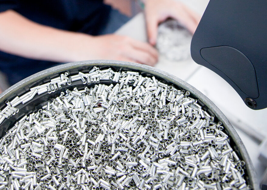 Nahaufnahme einer Schale mit zahlreichen kleinen, silberfarbenen Metallhülsen oder -rohren. Im Hintergrund sind unscharf die Hände einer Person bei einer Prüftätigkeit zu erkennen – die Szene steht für industrielle Fertigung und Qualitätssicherung von Präzisionsteilen.