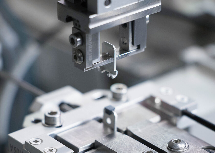 Detailaufnahme einer automatisierten Fertigungsstation, bei der ein metallisches Stanzbiegeteil von einem Greifer exakt positioniert wird. Die Szene zeigt präzise Handhabung in der industriellen Montage oder Qualitätssicherung von Verbindungselementen.