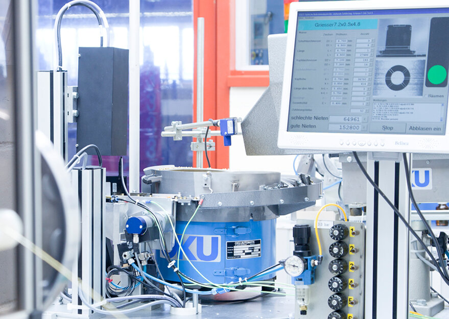 Automatisierte Prüfanlage mit Rundförderer und angeschlossenem Monitor, der Prüfdaten und Auswertungen für Kleinteile anzeigt. Die industrielle Anlage dient der Qualitätskontrolle, Sortierung und Zählung, etwa von Verbindungselementen wie Rohrnieten oder Stanzteilen.