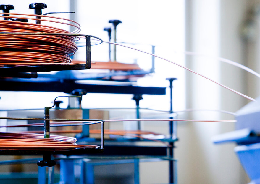 Mehrere aufgewickelte Kupferdrahtrollen auf Abrollgestellen in einer Produktionshalle. Die Drähte werden kontrolliert abgeführt und für die Weiterverarbeitung in automatisierten Drahtbearbeitungsmaschinen vorbereitet.