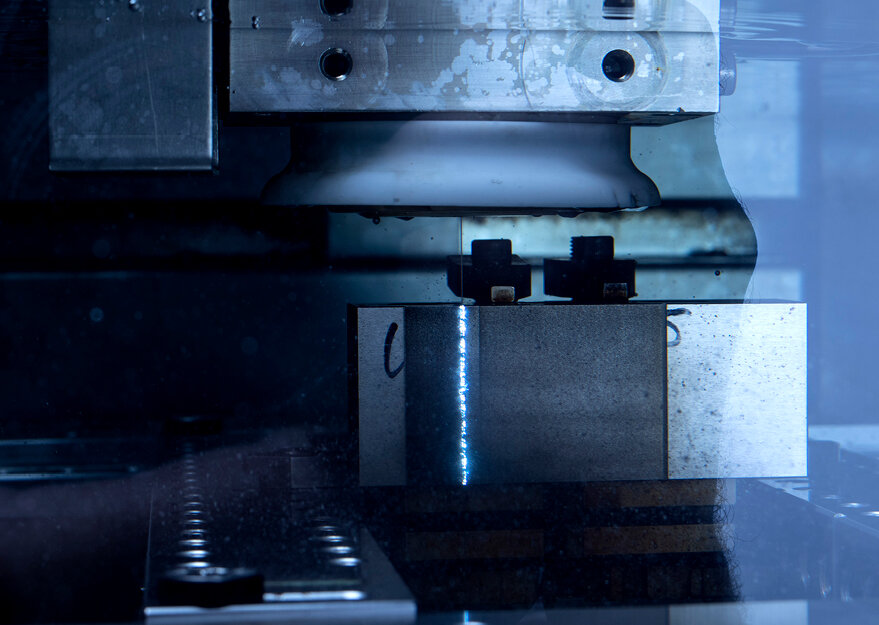 Detailaufnahme einer Drahterodiermaschine, bei der ein Werkstück aus Metall präzise bearbeitet wird. Ein feiner Lichtbogen ist entlang der Schnittkante zu sehen, was auf den laufenden Erodierprozess hinweist.