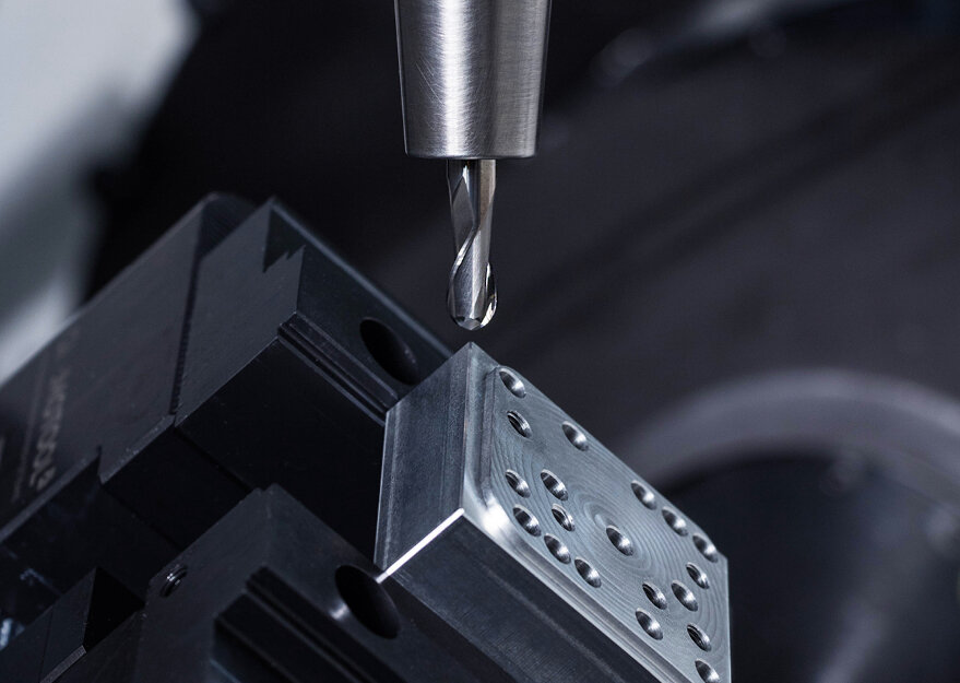 Nahaufnahme einer 5-achsigen CNC-Fräsmaschine beim Bearbeiten eines rechteckigen Metallblocks mit mehreren Bohrungen. Der Fräskopf ist präzise über dem Werkstück positioniert und bereit zur Bearbeitung.