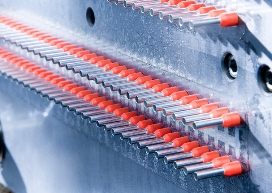 In einem Fertigungsprozess aufgereihte silberfarbene Aderendhülsen mit roten Isolierkragen, die präzise in mehreren Reihen aus einer Maschine austreten. Die Aufnahme zeigt die automatisierte Herstellung der Hülsen.