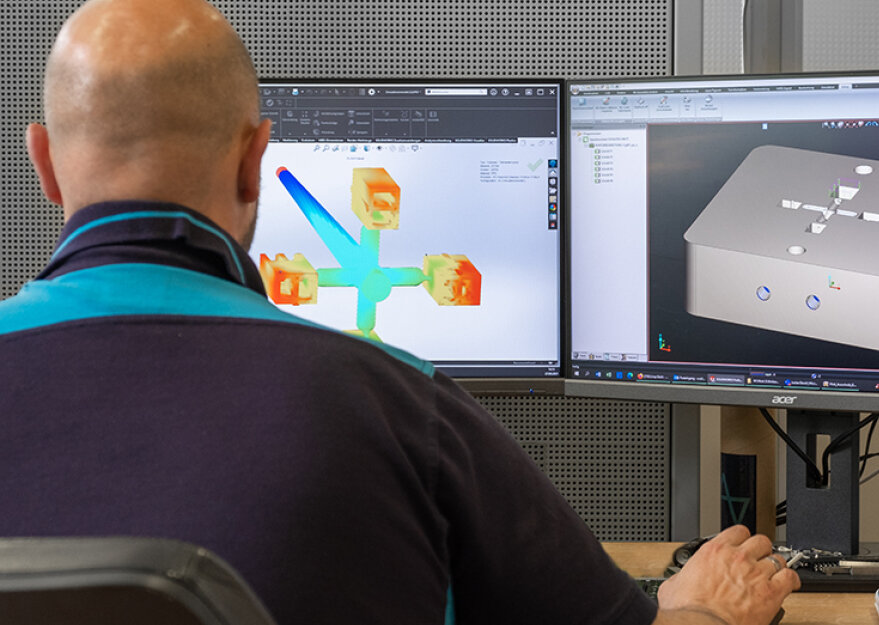 Ein Mitarbeiter betrachtet zwei Monitore mit CAD-Software, auf einem ist eine Mold-Flow-Simulation mit farbigen Flussanalysen zu sehen, auf dem anderen ein 3D-Modell eines Bauteils. Die Szene zeigt die digitale Planung und Analyse im Kunststoffspritzguss.
