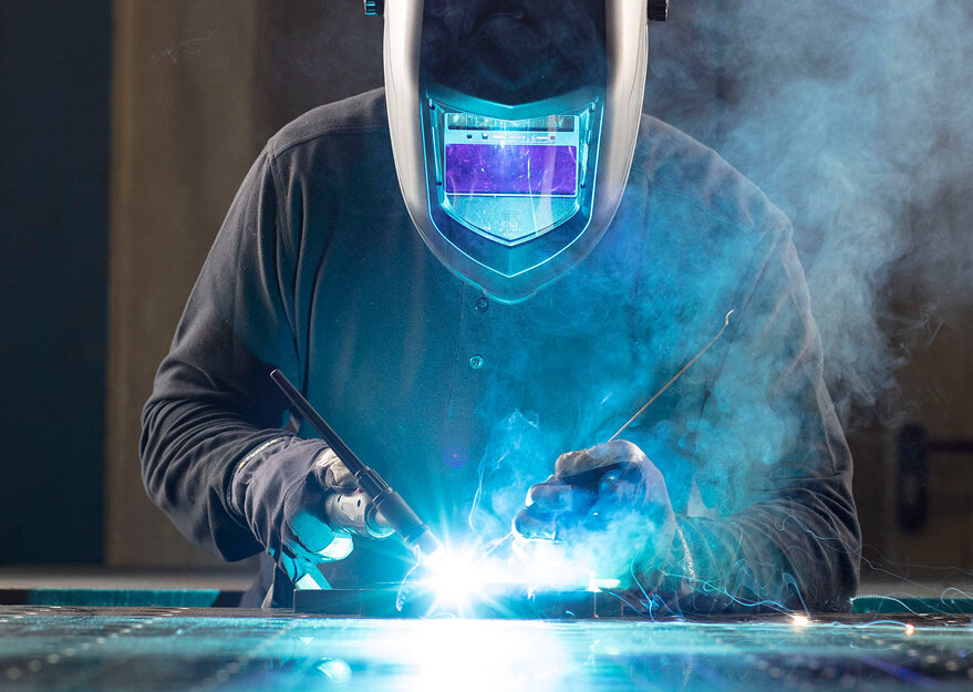 Eine Person mit Schweißhelm und Schutzhandschuhen beim Schweissen von Metall, umgeben von leuchtendem Lichtbogen und Rauch. Die Szene zeigt einen intensiven Moment aus dem Fertigungsprozess.