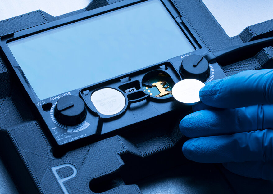 Eine Hand mit blauen Handschuhen setzt eine Knopfzelle in ein elektronisches Gerät mit großem Display ein. Das Gehäuse zeigt Beschriftungen wie „Sensitivity“ und „Delay“ sowie Drehregler zur Einstellung.