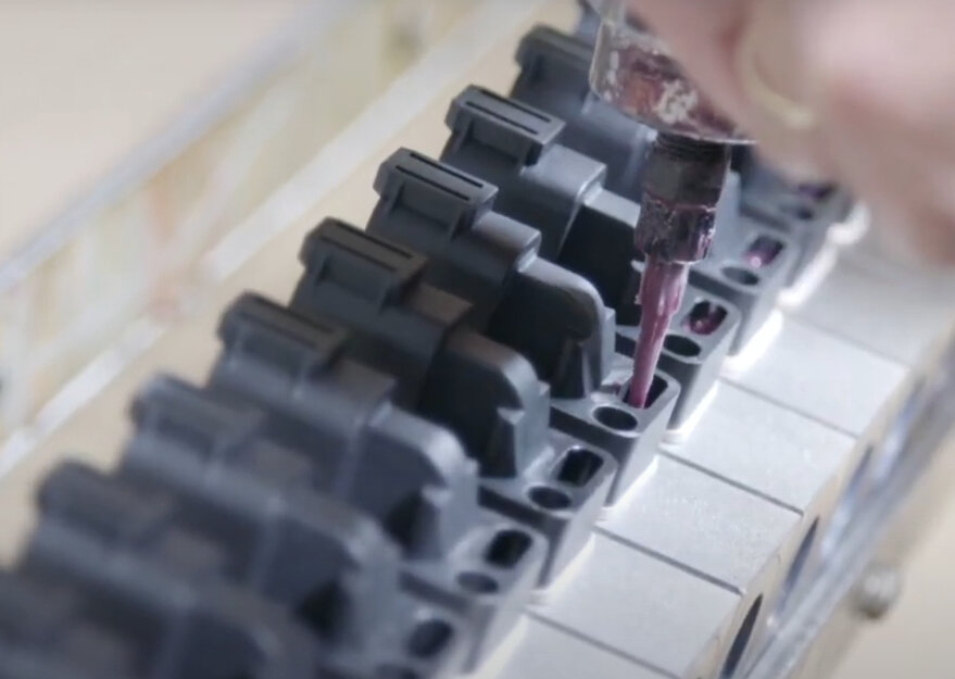 Nahaufnahme eines Fertigungsprozesses, bei dem schwarze Kunststoffgehäuse von Hand mit einer violetten Flüssigkeit befüllt werden. Die Bauteile sind in einer Reihe auf einer Vorrichtung fixiert, während eine Person mit einem Applikator präzise arbeitet.