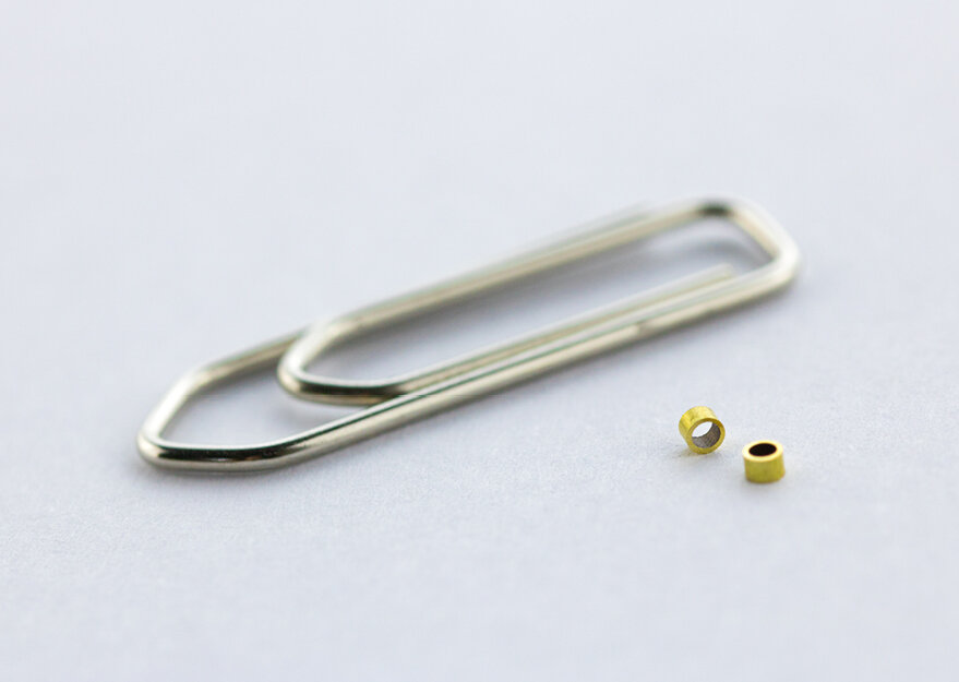 Zwei mikropräzise gefertigte Rohrabschnitte aus Messing mit sehr kleinem Durchmesser, abgebildet neben einer Büroklammer als Maßstabsreferenz auf neutralem Hintergrund. Die Geometrie deutet auf Anwendungen in der Feinmechanik oder Mikrosystemtechnik hin.