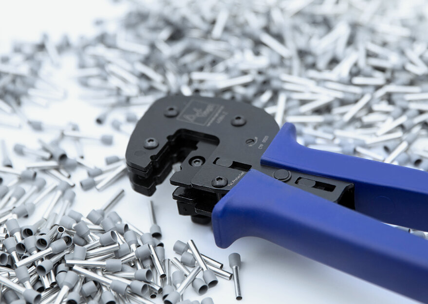 Crimpzange mit blauen Griffen liegt auf einer großen Menge isolierter Aderendhülsen aus Metall mit Kunststoffkragen. Das Werkzeug ist für das präzise Verpressen der Hülsen an Litzenleitungen in der elektrischen Verbindungstechnik ausgelegt.