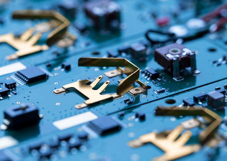 Nahaufnahme mehrerer Leiterplatten mit aufgelöteten vergoldeten Batteriehaltern aus gestanztem Metall, die zur Aufnahme von Knopfzellen dienen. Die Platinen sind mit verschiedenen SMD-Bauteilen bestückt und zeigen eine präzise Fertigung für den Einsatz in elektronischen Geräten.