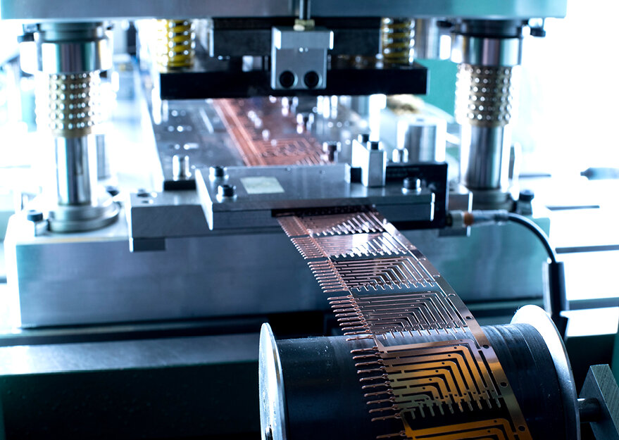 Präzisions-Stanzautomat im Einsatz bei der Verarbeitung eines endlosen Kupferbandes mit feinen Leiterstrukturen. Die Maschine führt das Material durch ein mehrstufiges Stanzwerkzeug, das für die Herstellung komplexer Stanzgitter in der Elektronikfertigung konzipiert ist.