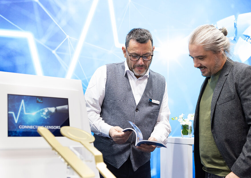 Zwei Männer im Gespräch auf einer Fachmesse, einer blättert in einer Broschüre, während der andere interessiert zuschaut; im Vordergrund ein Monitor mit der Aufschrift „Connected Sensors“ und stilisierter Handgrafik, im Hintergrund ein technisches, blaues Messewanddesign.