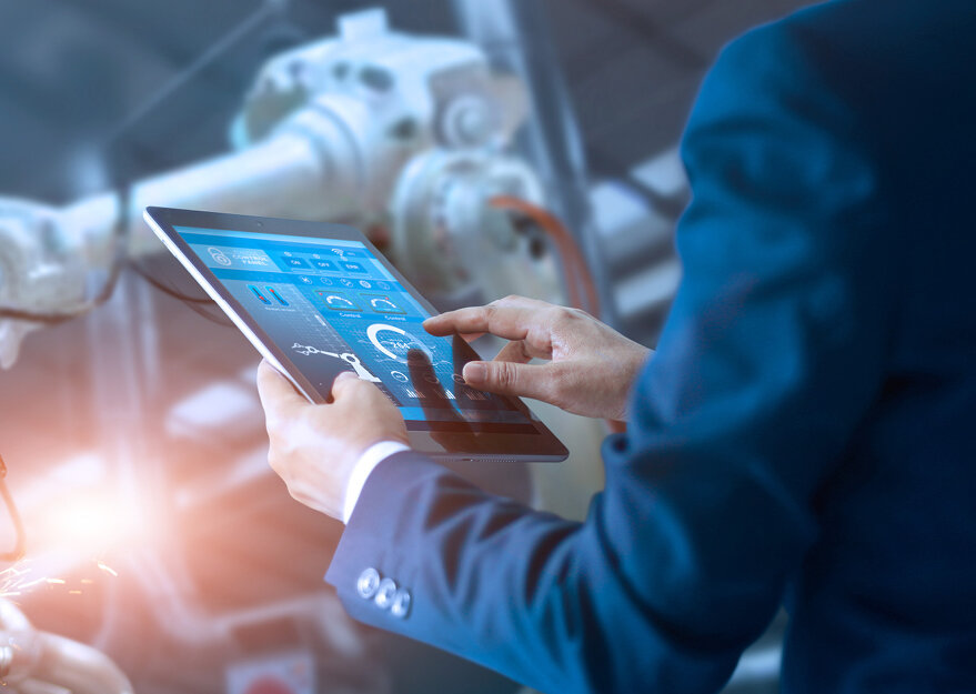 Mann in Anzug bedient ein Tablet mit digitaler Benutzeroberfläche zur Maschinensteuerung, im Hintergrund ist ein Industrieroboter in einer modernen Fertigungsumgebung zu sehen.