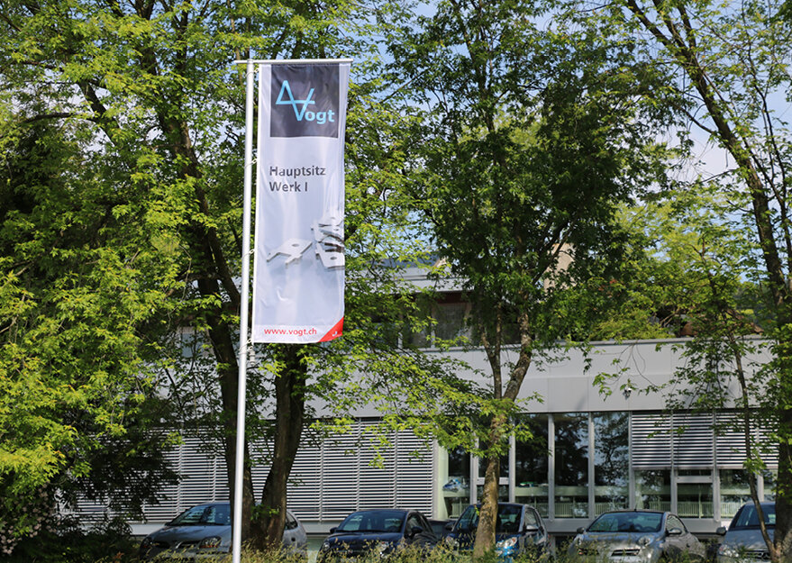 Fahne mit dem Logo der Vogt AG und der Aufschrift „Hauptsitz Werk I“ vor einem modernen Industriegebäude, umgeben von Bäumen und geparkten Autos.