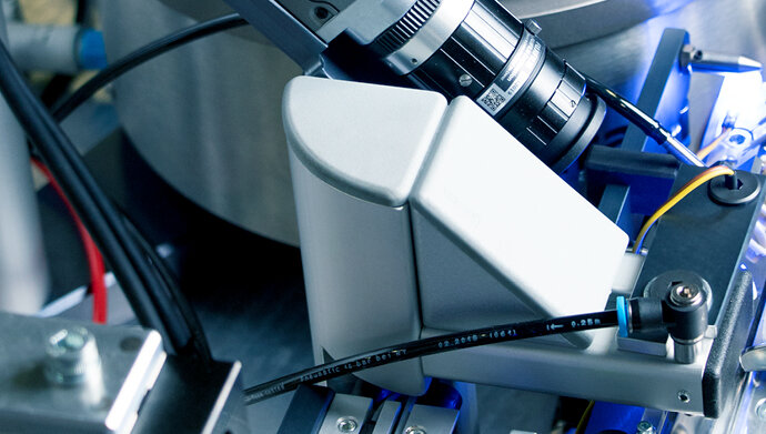 Nahaufnahme einer industriellen Kameraeinheit mit Fujinon-Objektiv, die über ein Förderband ausgerichtet ist und von Kabeln sowie einer Abdeckung umgeben ist. Die Szene zeigt einen Teil eines automatisierten Prüf- oder Sortiersystems mit präziser Beleuchtung.