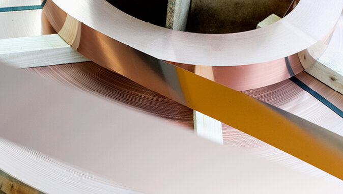 Close-up of a wound copper foil or metal strip roll with a shiny surface. The roll lies on a wooden holder and shows different colour reflections caused by the light.