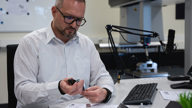 Ein Mann mit Brille und weißem Hemd sitzt an einem Schreibtisch und inspiziert ein technisches Bauteil mit einem Messgerät. Im Hintergrund ist ein Koordinatenmessgerät zu sehen, das auf präzise Qualitätsprüfung im Messlabor hinweist.