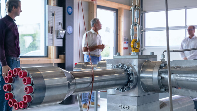 Große metallische Prüf- oder Fertigungseinrichtung mit mehreren Anschlüssen und rot markierten Öffnungen steht in einer Werkhalle, während drei Männer im Hintergrund das System betrachten. Die Szene wirkt wie eine Vorführung oder technische Besichtigung.