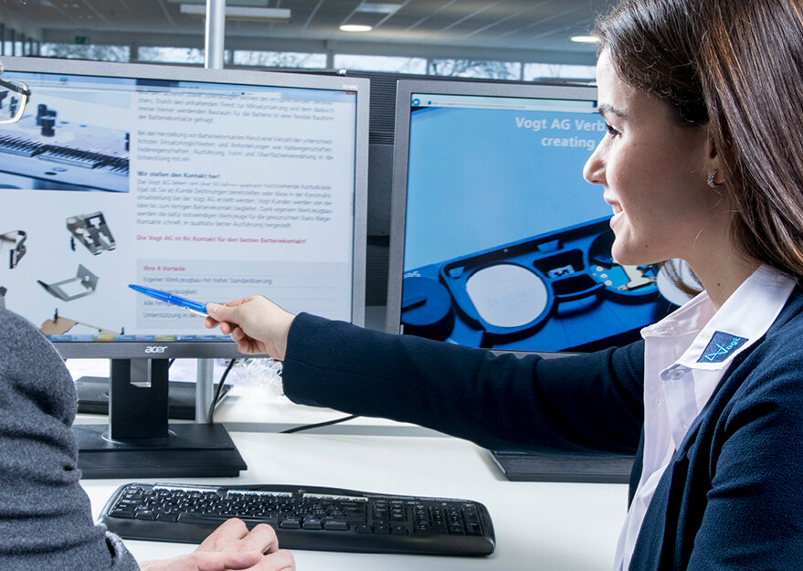Eine Mitarbeiterin im Business-Outfit zeigt mit einem Kugelschreiber auf Inhalte am Bildschirm und bespricht etwas mit einer weiteren Person. Im Hintergrund sind zwei Monitore mit technischen Informationen und einer Website der Vogt AG zu sehen – die Szene steht für Beratung, Kundenservice oder technische Verkaufsunterstützung.