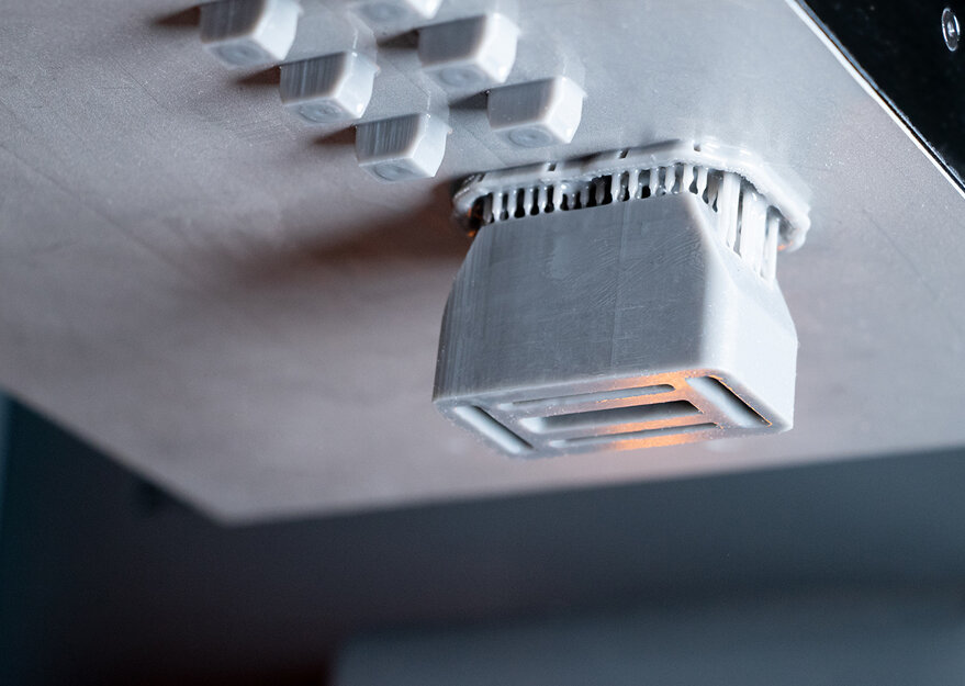 Ein Bauteil wird gerade kopfüber in einem 3D-Drucker gefertigt. Die einzelnen Schichten des Druckprozesses sind deutlich sichtbar, das Druckobjekt ist grau und besteht aus Kunststoff.