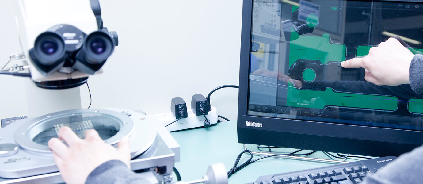 Eine Person bedient ein digitales Messsystem mit Mikroskop und zeigt auf den Monitor, der ein stark vergrössertes Bild eines technischen Bauteils anzeigt. Der Arbeitsplatz ist mit einem Computer, einem optischen Messgerät und mehreren Kabelverbindungen ausgestattet.