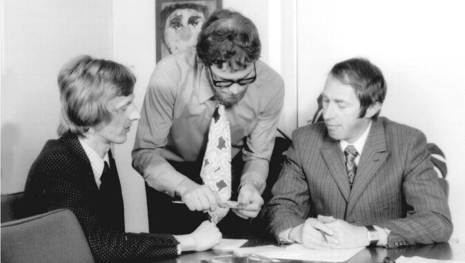 Schwarz-weiß-Fotografie aus den 1970er-Jahren, die drei Männer im Anzug an einem Besprechungstisch zeigt. Einer der Männer steht und erklärt den beiden sitzenden Kollegen etwas anhand eines Gegenstandes, vermutlich im Rahmen eines technischen oder geschäftlichen Gesprächs.